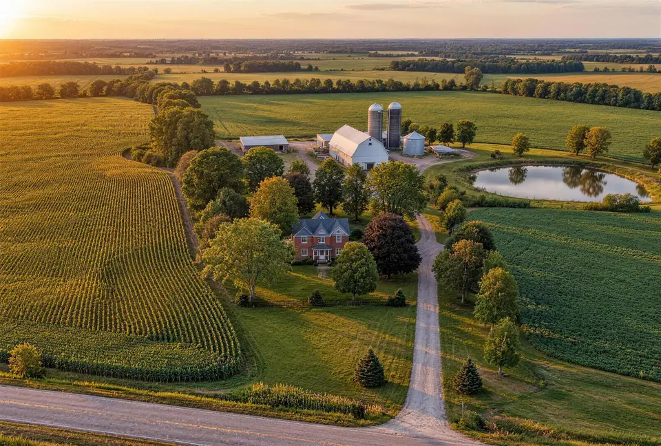 Aerial view of a farmhouse, barns and silos among crop fields, with a long lane and a pond at sunset